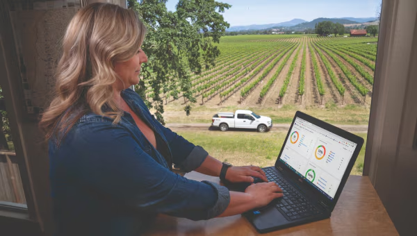 Woman working on laptop with fleet dashboard near vineyard and parked Ford truck.
